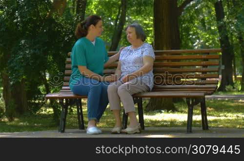 Young nurse spending sunny day with senior woman talking on park bench. Full HD footage in slow motion.