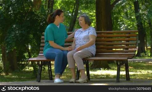 Young nurse spending sunny day with senior woman talking on park bench. Full HD footage in slow motion.