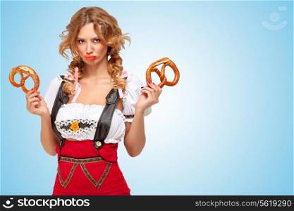 Young offended sexy Swiss woman wearing red jumper shorts with suspenders in a form of a traditional dirndl, holding two pretzels on blue background.