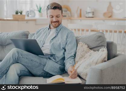 Young office worker working remotely from home, researching in internet using notebook, writing information down into red note book with pencil while sitting on cozy couch. Young office worker working remotely from home researching using notebook and taking notes