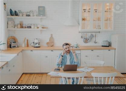 Young office worker working remotely from home with computer, sitting by kitchen table, tasked with important project, trying to find out solution, writing down and taking notes. Young office worker working remotely from home with computer while sitting by kitchen table