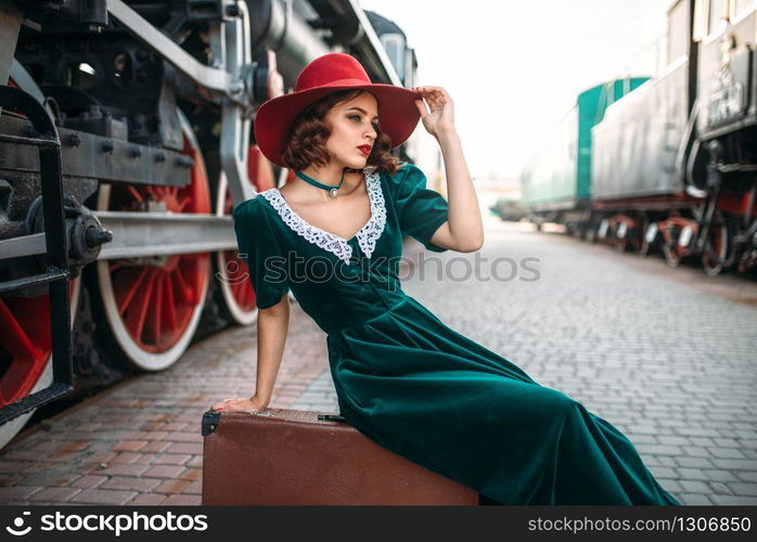 Young old-fashioned woman sitting on suitcase against vintage steam train, red wheels closeup. Old locomotive. Railway engine, travel by railroad. Woman sitting on suitcase against steam train