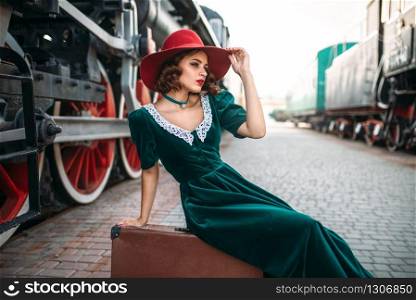 Young old-fashioned woman sitting on suitcase against vintage steam train, red wheels closeup. Old locomotive. Railway engine, travel by railroad. Woman sitting on suitcase against steam train