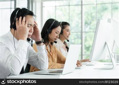 Young operator man headache while working with headset doing customer support at work. Group of Asian employee work in telemarketing customer service teams. Call Center with problem concept