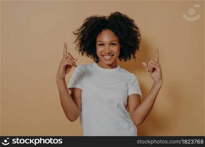 Young overjoyed excited dark skinned woman with curly hair wearing white tshirt pointing up with forefingers and showing blank copy space for advertising, isolated on beige studio background. Young overjoyed dark skinned woman pointing up with forefingers, isolated on beige background