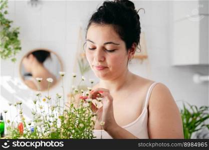 Young overweigh woman looking at chamomile flowers in white bathroom. Natural cosmetics concept. Body posititvity. Young overweigh woman looking at chamomile flowers in bathroom