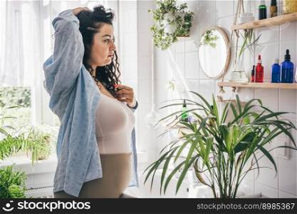 Young overweight brunette woman looking into mirrow in light eco friendly bathroom with biophilic design. Wellness, self confidence and body positivity concept. Young overweight brunette woman looking into mirrow