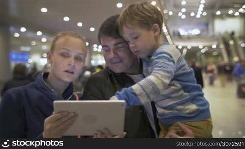 Young parents and little son at the crowded airport. Father holding boy who playing on tablet computer. Mom and dad discussing where to go and then they all leaving