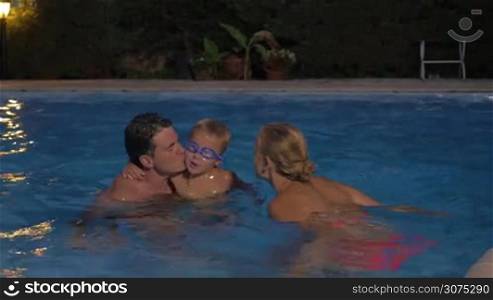 Young parents and little son bathing in outdoor pool in the evening. They kissing each other and cheerful boy splashing water