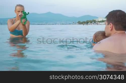 Young parents and little son bathing in the sea. Mom and child playing with windup frog. Family summer vacation