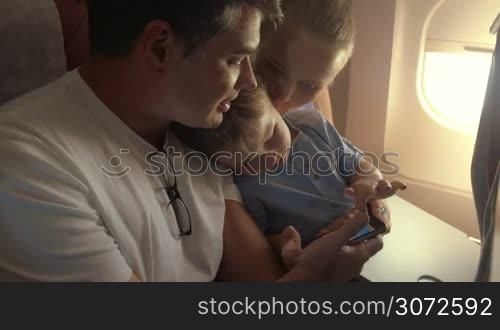 Young parents and little son traveling by air. Child playing game on fathers smart phone to entertain himself during the flight. Mother kissing the child