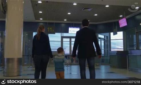 Young parents and little son walking in airport terminal. They coming up to the glass door and looking out