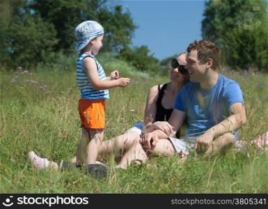 Young parents and son on the meadow on a hot summer day. Happy couple sitting on the grass and looking at the kid. Outdoor family activity