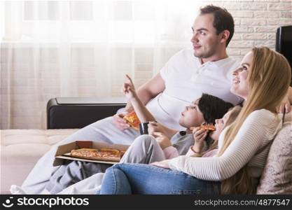 Young parents and their children are watching TV, eating pizza and smiling while sitting on couch at home