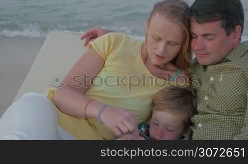 Young parents and their son are resting in lounge chair by the sea. Boy is playing with smartphone.