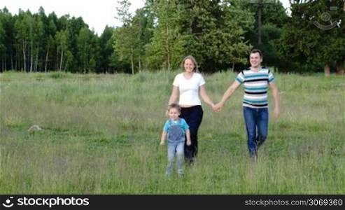 Young parents and their son walking on the meadow. Man and woman holding hands. Wood in background
