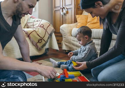 Young parents playing with their toddler son a wooden game building in the living room. Parents playing with toddler a wooden game building