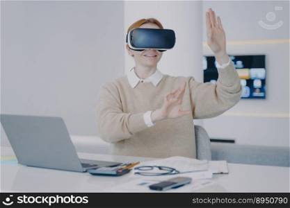 Young€pean woman in VR headset in office. Girl is sitting at the desk in 3d gogg≤s in front of laptop. Working on visual effects project in cyberspace. Concept of modern devices using.. Girl is sitting at the desk in 3d gogg≤s in front of laptop. Concept of modern devices using.