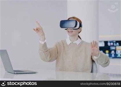 Young€pean woman in VR headset in office. Girl is sitting at the desk in front of laptop. Entrepre≠ur working on visual effects project in cyberspace. Concept of digital solutions in remote work.. Young€pean woman entrepre≠ur in VR headset. Concept of digital solutions in remote work.