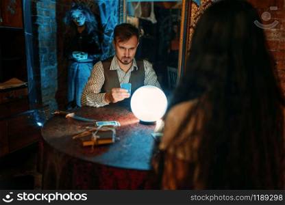 Young people at the table with crystal ball on spiritual seance, scary witch on background. Female foreteller calls the spirits. Young people at the table with crystal ball