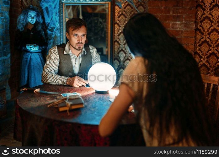 Young people at the table with crystal ball on spiritual seance, scary witch on background. Female foreteller calls the spirits