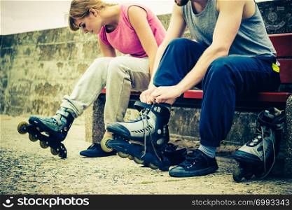Young people friends in training suit putting on roller skates outdoor. Woman and man sitting on bench.. People friends putting on roller skates outdoor.