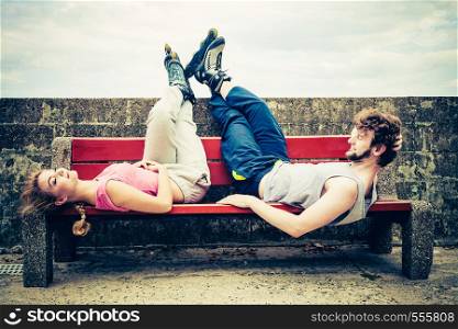 Young people friends in training suit with roller skates. Woman and man relaxing lying on bench outdoor.. Young people friends relaxing on bench.