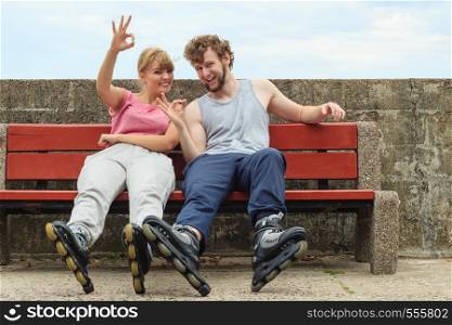 Young people friends in training suit with roller skates giving showing ok gesture. Woman and man relaxing on bench outdoor.. Young people friends relaxing on bench.