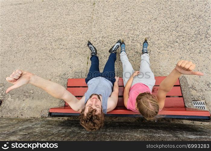 Young people friends in training suit with roller skates giving thumb up gesture. Woman and man relaxing on bench outdoor.. Young people friends relaxing on bench.