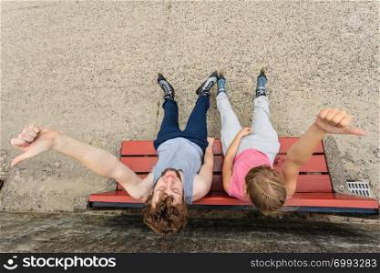 Young people friends in training suit with roller skates giving thumb up gesture. Woman and man relaxing on bench outdoor.. Young people friends relaxing on bench.