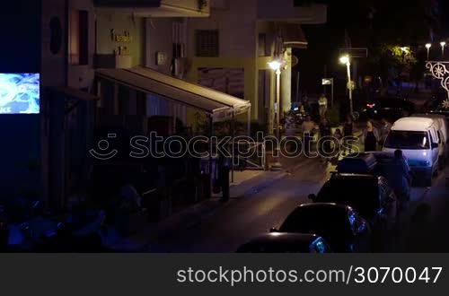 Young people walking along narrow street with shops and parked cars at night