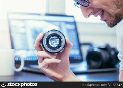 Young photographer is looking at a photography lens, laptop in the blurry background