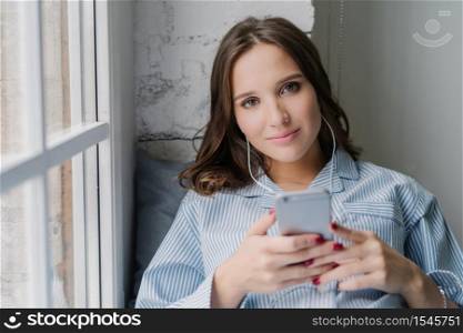Young pleasant looking woman satisfied with good sound in earphones, holds modernn cell phone, dressed casually, enjoys leisure time, sits on window sill. Female listens playlist songs indoor