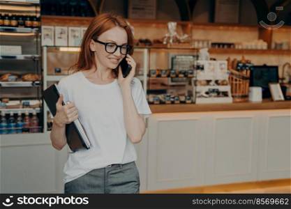 Young pleasant redhead woman freelancer talking on phone and smiling while leaving cafe or coffee shop, business woman in casual clothes with laptop using cellphone on city street outdoors. Young pleasant redhead woman freelancer talking on phone and smiling while leaving cafe coffee shop