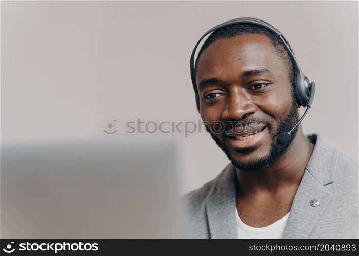 Young pleased african american man office worker in headset communicating with colleagues during video call on laptop, smiling guy call center agent talking with client, consulting customer online. Young pleased african american man callcenter agent talking with client, consulting customer online