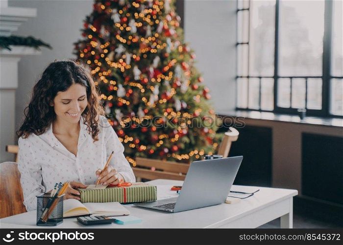 Young pleased european woman office worker writing christmas letter, greeting card with holiday wishes while sitting at her workplace with laptop and working remotely from home during winter holidays. Pleased european woman office worker writing christmas letter, greeting card with holiday wishes