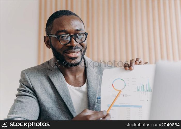 Young positive african man employee sits at desk having video conference online with partner, holds accounting report in hand and points to calculations. Afro american business man during videcall. Young positive african man employee sits at desk having video conference online with partner