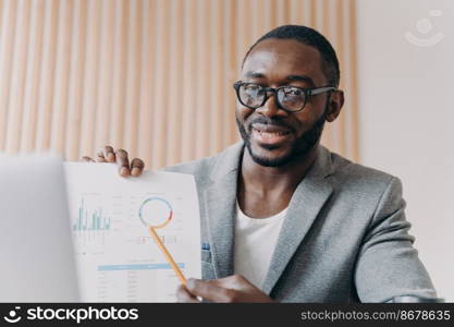 Young positive african man employee sits at desk having video conference online with partner, holds accounting report in hand and points to calculations. Afro american business man during videcall. Young positive african man employee sits at desk having video conference online with partner