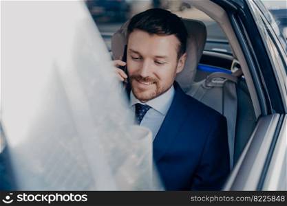 Young positive businessman rides in car on passenger seat to office in city center, talking on smartphone with focused look and smiling. Side image of entrepreneur in suit reading newspaper in auto. Businessman rides in black car in passenger seat and talking on cellphone