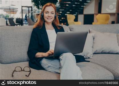 Young positive gingerhair lady freelancer, female helpline operator with wireless headphones and laptop, working remotely online while sitting on cozy sofa in coffee shop, looking at camera with smile. Young positive gingerhair lady freelancer helpline operator with wireless headphones and laptop