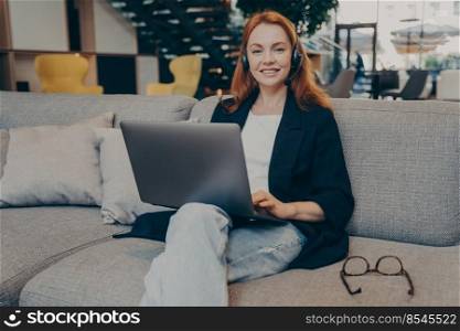 Young positive gingerhair lady freelancer, female helpline operator with wireless headphones and laptop, working remotely online while sitting on cozy sofa in coffee shop, looking at camera with smile. Young positive gingerhair lady freelancer helpline operator with wireless headphones and laptop