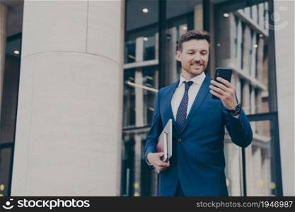 Young positive handsome businessman in elegant outfit using mobile phone, texting sms or checking email while standing outdoors against office building after hard working day, holding laptop in hand. Young handsome businessman using mobile phone, texting sms or checking email, standing outdoors