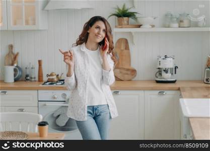 Young positive hispanic woman talking on phone in her stylish kitchen at home. Relaxed pretty housewife. Modern white scandinavian interior. Stove, worktop and cuisine.. Young hispanic woman talking on phone. Relaxed housewife in her stylish kitchen at home.