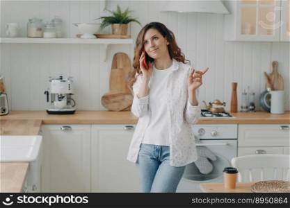 Young positive hispanic woman talking on phone in her stylish kitchen at home. Relaxed pretty housewife. Modern white scandinavian interior. Stove, worktop and cuisine.. Young hispanic woman talking on phone. Relaxed housewife in her stylish kitchen at home.