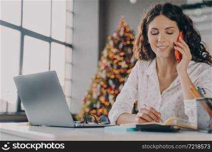 Young positive Italian woman with smartphone doing online shopping on laptop during Christmas holidays, smiling female freelancer talking on mobile while woking from home office in room with xmas tree. Positive Italian woman with smartphone doing online shopping on laptop during Christmas holidays