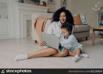 Young positive mixed race mother and cute boy son having video call with relatives, father or grandparents, sitting with laptop on floor and smiling, child wearing headset and pointing at screen. Young positive mixed race mother and cute boy son having video call on laptop