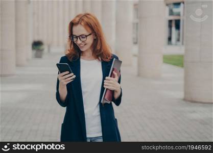 Young positive red haired business woman in elegant outfit using mobile phone, texting sms or checking email while standing outdoors after hard working dy, holding laptop and note book. Young positive red haired business woman in elegant outfit using mobile phone, standing outdoors