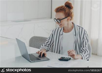Young positive red-haired woman accounter or financier working remotely on laptop computer and using calculator while sitting at her home office, smiling while looking at pc screen, dressed casually. Young positive red-haired woman accounter working remotely on laptop and using calculator