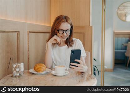 Young positive redhead woman in earphones sitting in cafe and talks online on smartphone, female blogger records video for social account or her blog, smiles and communicates with followers. Young positive redhead woman in earphones sitting in cafe and talks online on smartphone