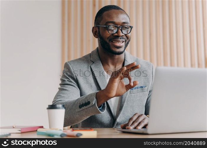 Young positive sitisfied african american businessman in suit talking with business partner online, showing okay gesture during video call on laptop computer, taking part in web conference. Young positive sitisfied african american businessman in suit talking with business partner online